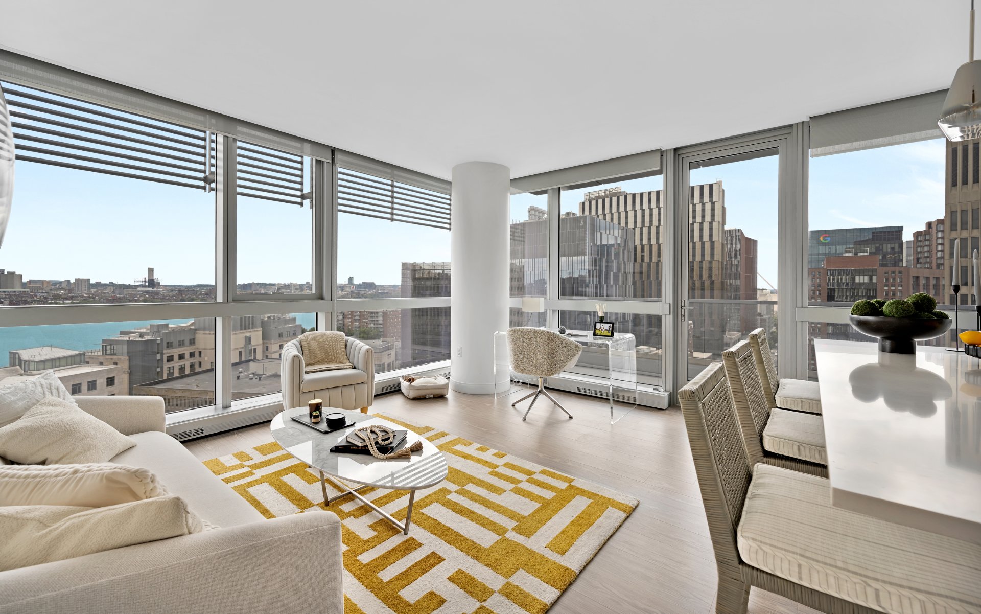 White-themed living area with floor-to-ceiling glass windows showcasing the city skyline. Warm brown wooden flooring accented by a yellow carpet
